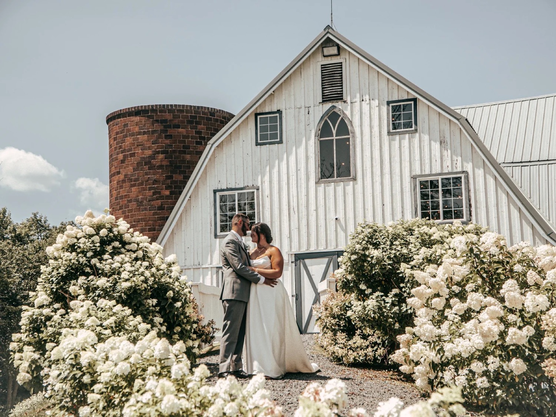 Newlyweds embracing beside white hydrangeas at a white barn estate